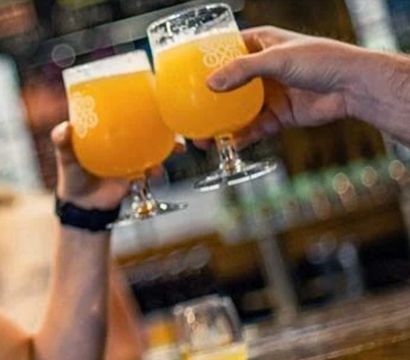 Two people clinking tulip glasses of hazy orange craft beer at a lively taproom — casual cheers