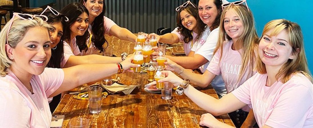 Group of eight women in matching pink shirts clinking small craft-beer glasses over a rustic wooden table in a bright taproom, celebrating together.