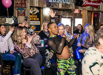 Energetic dancer in neon outfit giving a playful lap dance to a laughing girls' night crowd inside a lively country-style bar with neon beer signs and phones raised to record.