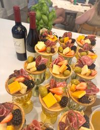 Mini charcuterie cups on glass goblets with cubes of cheddar and mixed cheeses, folded salami, green olives, blackberries and strawberry halves on wooden rounds, set beside wine bottles and a basil plant — elegant appetizer display.