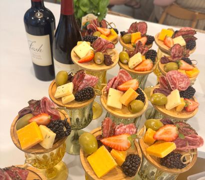 Mini charcuterie cups on glass goblets with cubes of cheddar and mixed cheeses, folded salami, green olives, blackberries and strawberry halves on wooden rounds, set beside wine bottles and a basil plant — elegant appetizer display.