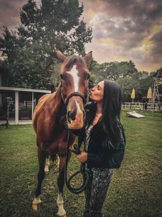 Woman kissing a chestnut horse with a white blaze while holding its lead rope in a grassy paddock under a dramatic cloudy sunset sky near trees and stables.