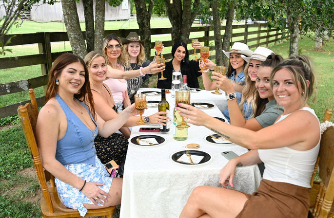 Group of women toasting with wine glasses at a long white-tablecloth outdoor brunch under trees by a wooden fence on a country lawn, cheerful rustic gathering.