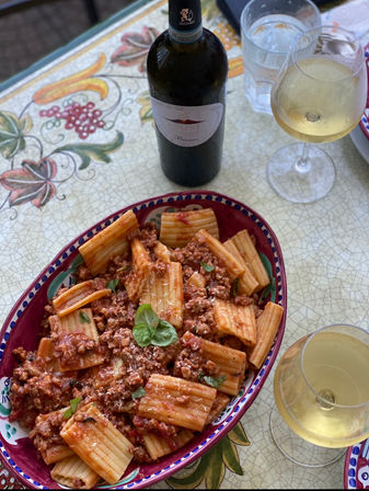 Hearty rigatoni in tomato-meat ragù served in a colorful ceramic bowl on a hand-painted tiled table, paired with a bottle and two glasses of chilled white wine