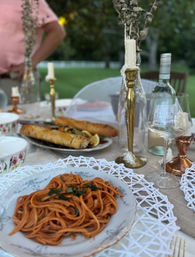 Al fresco garden dinner table with a plate of tomato spaghetti on vintage china, crusty baguette on a platter, melted-candle brass candlesticks, wine bottle and glass on a rustic outdoor setting.