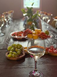 Rustic charcuterie spread on a wooden table: green grapes on a board, cubes of cheese and sliced salami on a raised plate, and a white wine glass with a blurred centerpiece and glasses in the background