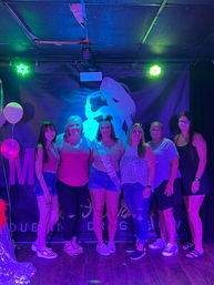 Six women posing on a neon-lit club stage for a bachelorette party photo; center woman wears a sash and denim shorts, purple and green lights illuminate balloons and a silhouetted backdrop.