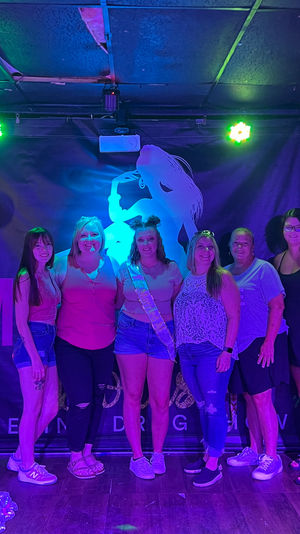 Six women posing on a neon-lit club stage for a bachelorette party photo; center woman wears a sash and denim shorts, purple and green lights illuminate balloons and a silhouetted backdrop.