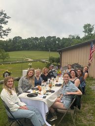 Group of women enjoying an outdoor farmhouse dinner at a long, flower-adorned table with amber goblets by a barn and American flag, set against rolling green fields, trees, string lights, and an overcast sky.