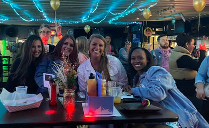 Four friends smiling at a high-top table in a neon-lit bar with blue string lights and gold balloons, drinks, condiments and a small flower vase on the table, other patrons mingling in the background.