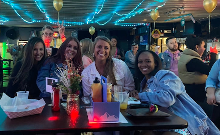 Four friends smiling at a high-top table in a neon-lit bar with blue string lights and gold balloons, drinks, condiments and a small flower vase on the table, other patrons mingling in the background.