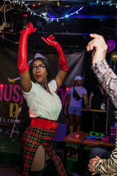 Drag performer in red opera gloves, white blouse and plaid skirt striking a dramatic pose on a crowded nightclub stage under string lights.