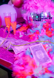 Neon pink nightclub party table with a mirrored disco ball, colorful feather boas, scattered dollar bills and two champagne flutes under purple lighting
