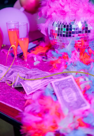 Neon pink nightclub party table with a mirrored disco ball, colorful feather boas, scattered dollar bills and two champagne flutes under purple lighting