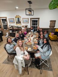Group of women enjoying a lively dinner party around a long wooden farmhouse table set with pasta, candles, wine and plates in a cozy rustic dining room with wood floors, pendant light and vintage decor.