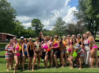 Group of women in colorful swimsuits and leis posing at a sunny countryside ranch by a wooden fence with a brown horse and red barn under a partly cloudy sky.