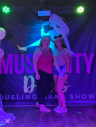 Two women smiling and posing in front of a neon-lit indoor stage backdrop with a dancer silhouette; one wears a celebratory sash and shorts, colorful stage lights and balloons create a lively party photo vibe.