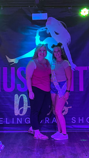 Two women smiling and posing in front of a neon-lit indoor stage backdrop with a dancer silhouette; one wears a celebratory sash and shorts, colorful stage lights and balloons create a lively party photo vibe.