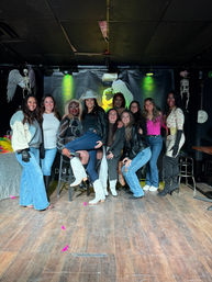 Group of friends and drag performers posing on a small nightclub stage under green spotlights, skeleton decorations on the sides, wooden floor, and a guest wearing a “bride” cowboy hat — fun nightlife group photo.