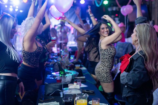 Women dancing and celebrating at a lively nightclub table with pink balloons, colorful lights, drinks and leopard-print outfits — energetic girls' night out vibe.