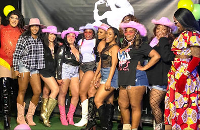 Vibrant group photo at a cowgirl-themed party: women and performers posing against a black backdrop with balloons, wearing pink cowboy hats, denim shorts, fishnet tights and colorful boots.