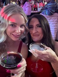 Two smiling women in red outfits toasting with dark espresso cocktails in coupe glasses during a festive night out at a lively bar with colorful lights and patrons in the background.