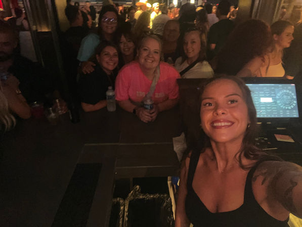 Smiling woman taking a selfie with a group of friends at a crowded indoor bar, people gathered around the bar counter under warm dim lights for a lively nightlife scene.