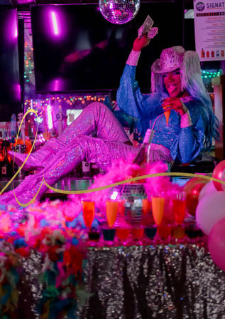 Drag performer in a sparkly blue outfit and silver fringe cowboy hat lounges on a neon-lit nightclub bar counter, waving cash and holding a cocktail under a disco ball with colorful drinks, balloons, and feather boas.