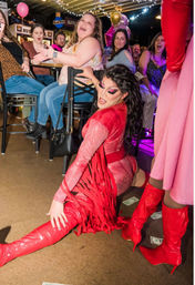 Drag performer in a sparkling red fringe outfit and glossy thigh-high boots doing a dramatic split on a nightclub floor while a lively seated audience cheers, claps and tips with dollar bills under string lights and balloons.