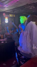 Bride-to-be in a veil at a lively nightclub bachelorette party, crowd cheering as a performer with a neon green wig entertains under colorful LED lights, drinks and dollar bills scattered on the table and floor.