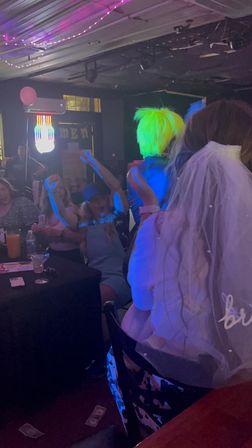 Bride-to-be in a veil at a lively nightclub bachelorette party, crowd cheering as a performer with a neon green wig entertains under colorful LED lights, drinks and dollar bills scattered on the table and floor.