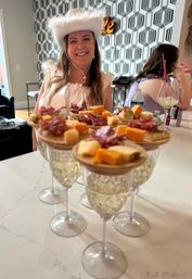 Smiling woman in a white feathered cowboy hat behind a row of charcuterie bites served on wine glasses at a marble kitchen island, modern geometric wallpaper and casual party scene