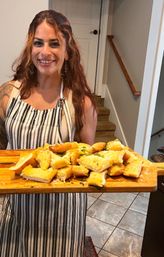 Smiling person in a striped apron holding a wooden serving board piled with freshly baked garlic bread garnished with parsley in a bright home kitchen with tiled floor and a staircase.
