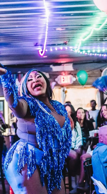 Drag performer in blue fringe costume waving a hand fan in a neon-lit crowded bar, audience cheering under string lights and balloons during a lively drag show.