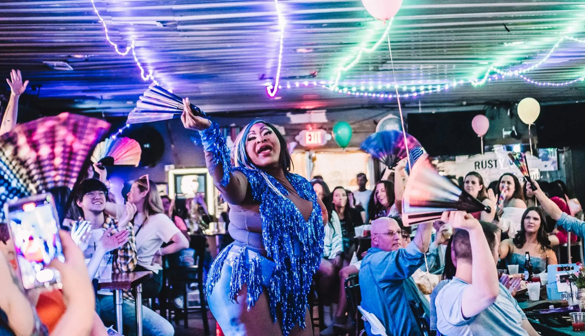 Drag performer in blue fringe costume waving a hand fan in a neon-lit crowded bar, audience cheering under string lights and balloons during a lively drag show.