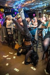 Performer in a rhinestone-studded black bodysuit and glittery silver boots doing a headstand on a cash-strewn bar floor while an excited crowd with phones watches under string lights and balloons.