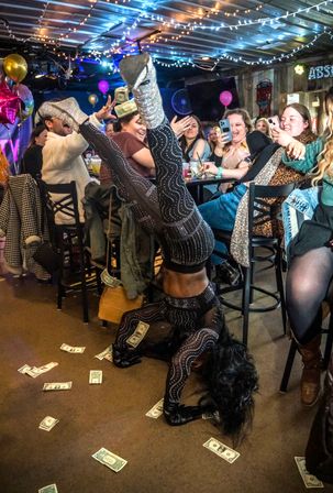 Performer in a rhinestone-studded black bodysuit and glittery silver boots doing a headstand on a cash-strewn bar floor while an excited crowd with phones watches under string lights and balloons.