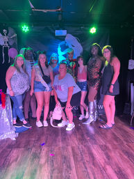 Group of friends posing with glammed-up drag performers on a nightclub stage under green and purple lights, one guest wearing a bachelorette sash, glittery costumes, silver boots, balloons and a skeleton decoration against a silhouette backdrop.