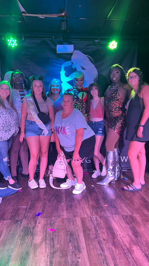Group of friends posing with glammed-up drag performers on a nightclub stage under green and purple lights, one guest wearing a bachelorette sash, glittery costumes, silver boots, balloons and a skeleton decoration against a silhouette backdrop.