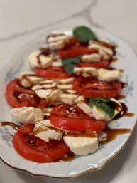 Close-up of a Caprese-style salad: sliced ripe red tomatoes layered with fresh mozzarella rounds, basil leaves and drizzled balsamic glaze on a white decorative platter