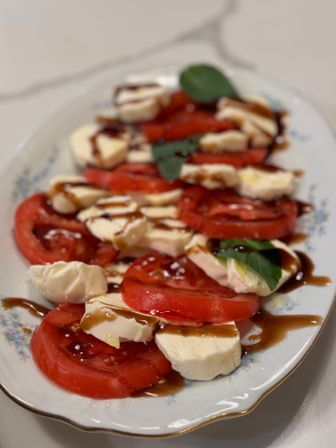 Close-up of a Caprese-style salad: sliced ripe red tomatoes layered with fresh mozzarella rounds, basil leaves and drizzled balsamic glaze on a white decorative platter