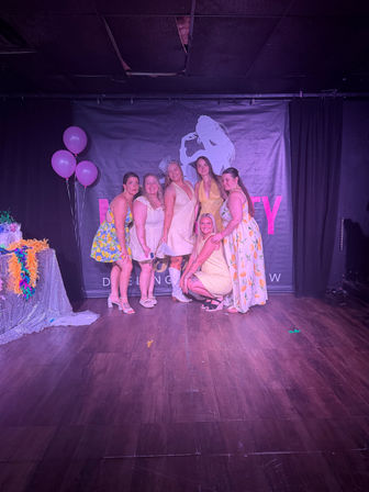 Six women in yellow and white dresses posing on an indoor stage under purple lighting with pink balloons and a singer-silhouette backdrop, festive party photo