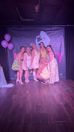 Six women in yellow and white dresses posing on an indoor stage under purple lighting with pink balloons and a singer-silhouette backdrop, festive party photo