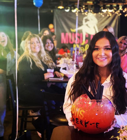 Smiling woman cradles an oversized red fishbowl cocktail with multiple straws at a lively indoor bar party, friends, balloons and a music banner in the background.