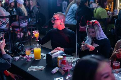Dimly lit bar scene with two people at a table — one in sunglasses animatedly raising an orange cocktail, the other with long blonde hair and red heart-shaped sunglasses on her head scrolling a phone; pitcher of orange juice, stacks of cash, condiments and glasses on the table, lively crowd in the background.