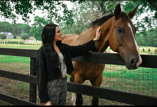 Smiling woman pets a curious brown horse over a wooden fence on a green country farm, with trees and rolling fields in the background — outdoor equestrian scene.