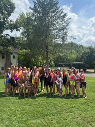 Cheerful group of women in colorful swimsuits and leis posing with a brown horse on a sunny green lawn in a rural backyard with a tall evergreen and farmhouse in the background — outdoor summer celebration.