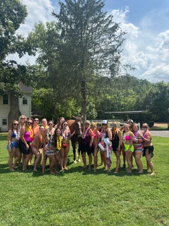 Cheerful group of women in colorful swimsuits and leis posing with a brown horse on a sunny green lawn in a rural backyard with a tall evergreen and farmhouse in the background — outdoor summer celebration.