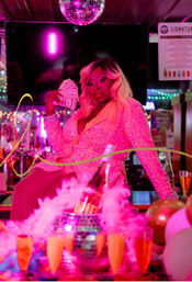 Drag performer in a sparkling pink sequined outfit holding fanned cash while posing on a bar counter under a disco ball, surrounded by pink neon lights, champagne bottles, balloons and party decorations in a nightclub.