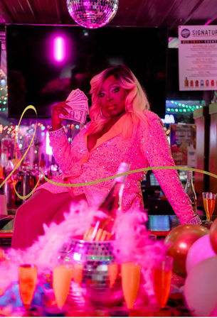 Drag performer in a sparkling pink sequined outfit holding fanned cash while posing on a bar counter under a disco ball, surrounded by pink neon lights, champagne bottles, balloons and party decorations in a nightclub.